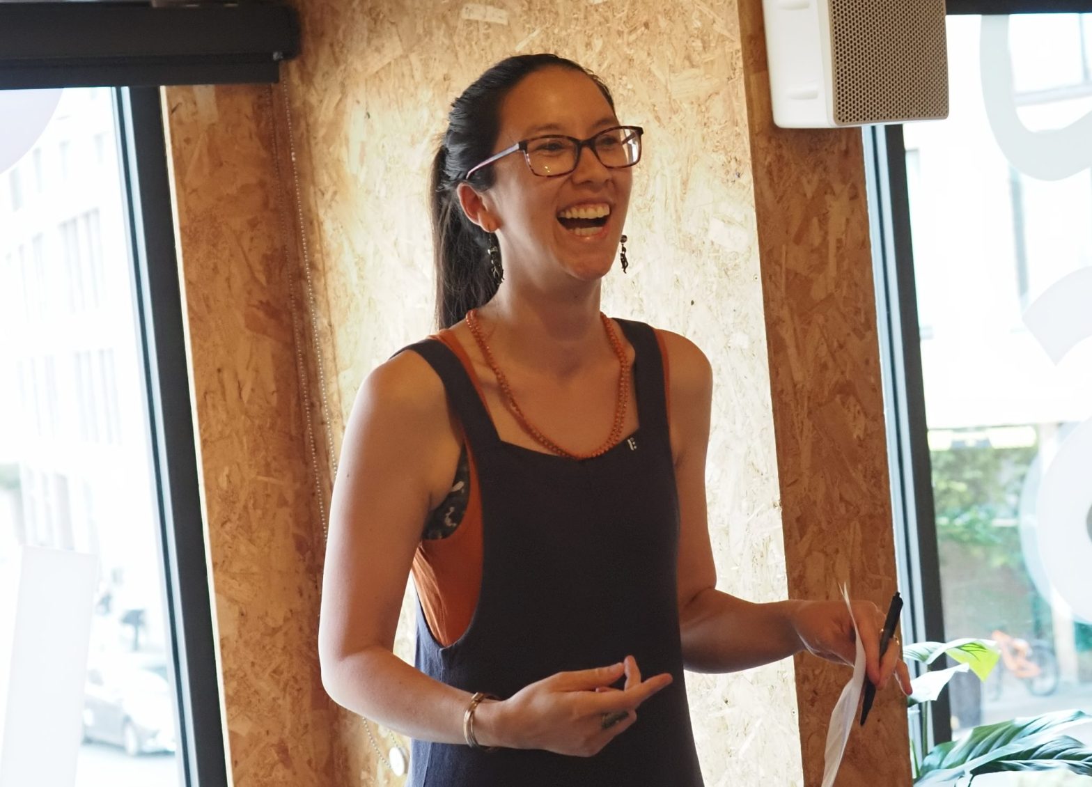 A woman stands in front of a window. She is smiling and holding a piece of paper