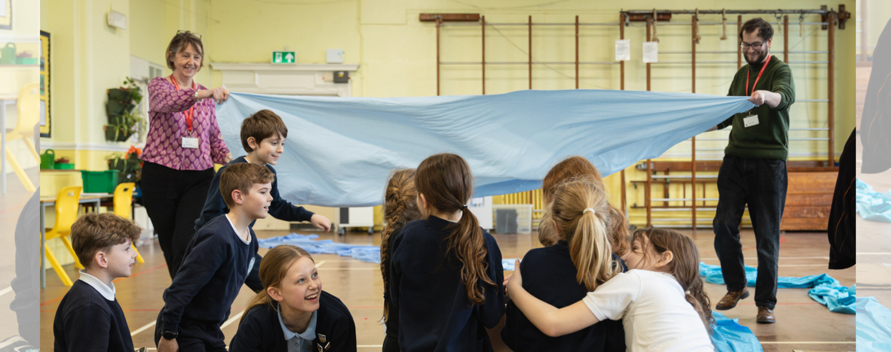 Two members of school staff (a man and a woman, wearing brightly coloured staff lanyards) gently lower a large piece of blue silk that represents a river onto the heads of the students sitting on the floor. The students are aged 8-10 and are smiling and laughing.